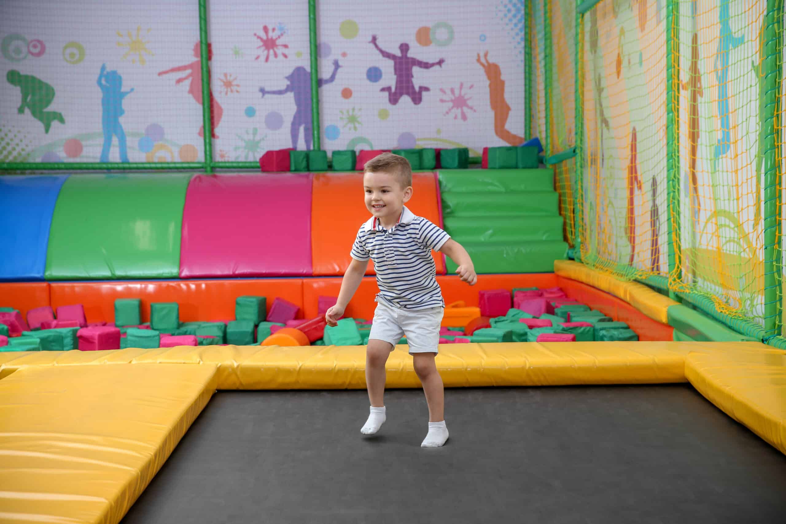 Jeune garçon joyeux en short et polo rayé sautant sur un trampoline dans une aire de jeux intérieure colorée avec murs décorés et coussins.