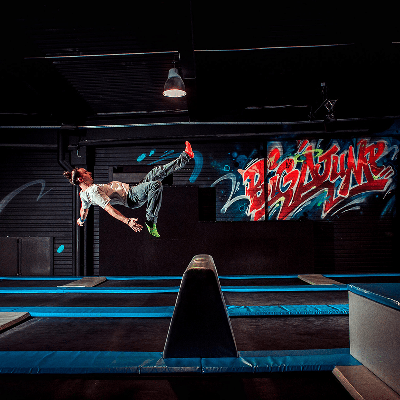 Homme en plein saut au-dessus de trampolines bleus, chaussettes vives. Graffiti "BIGJUMP" coloré en arrière-plan.