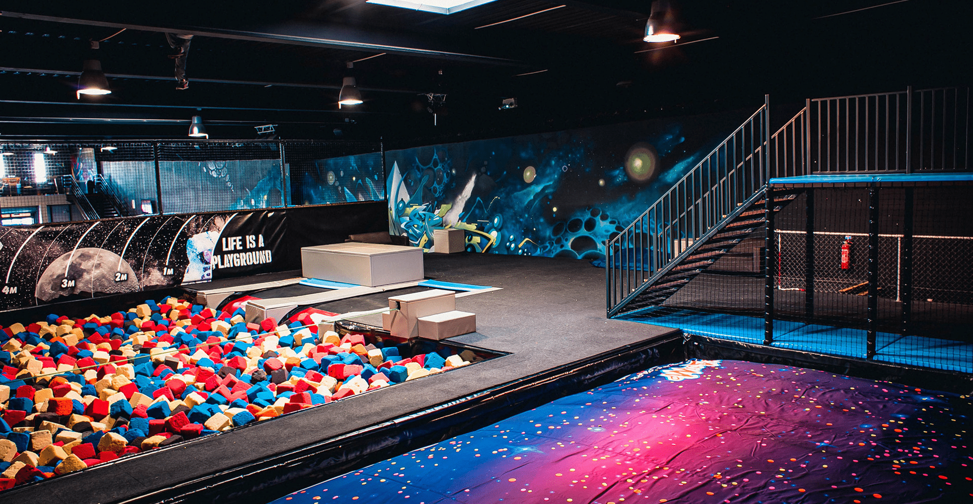 Trampoline park intérieur avec fosse à mousse colorée, trampolines spatiaux et murale galaxie. Texte: 'LIFE IS A PLAYGROUND'.