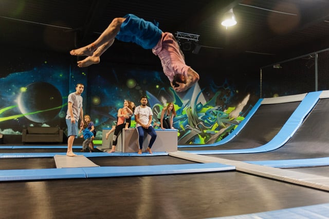 Trampoline Park: Salto, Fun et Acrobates en Intérieur Un homme réalise un salto arrière sur trampoline dans un parc indoor. Des adultes et enfants observent, les murs affichent des graffitis spatiaux.
